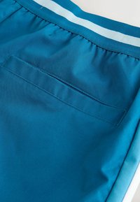 Détail de la poche arrière sur un short de sport bleu sarcelle avec une taille élastique ornée de bandes blanches et bleu sarcelle.
