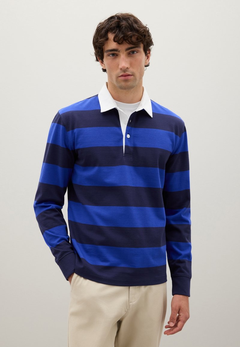 Mann in einem langärmeligen Rugby-Shirt mit breiten blauen und marineblauen Streifen, weißem Kragen und beigen Hosen, der vor einem einfarbigen hellen Hintergrund steht.