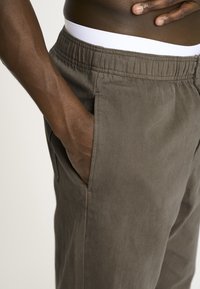 Mørk olivengrønne bukser med tekstureret stof, elastisk talje og sidelommer. Synlig syning og en tætsiddende pasform omkring taljen.