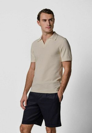 Polo a maniche corte, modello pullover, in tessuto leggermente beige chiaro texturizzato con colletto tradizionale, abbinata a pantaloncini scuri. Design semplice, linee pulite.
