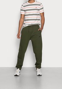 Gestreept wit T-shirt met rode en groene lijnen, gecombineerd met olijfgroene sweatpants met een elastische tailleband en zwart logo. Grijze sneakers.