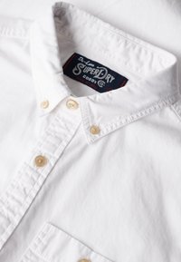 Camisa blanca de botón hecha de algodón con un cuello puntiagudo, que presenta botones beiges y una etiqueta que dice "SuperDry" en el interior del cuello.
