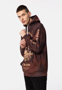 Brun tie-dye hoodie med vita dragsnören, har grafik och text i ljusbrunt. Ribbad mudd och detalj med framficka.