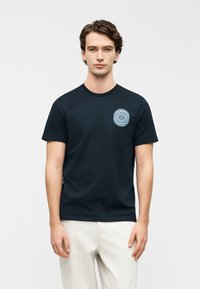 Μπλε βαμβακερό μπλουζάκι με στρογγυλή λαιμόκοψη και κοντά μανίκια, με ένα ανοιχτό μπλε κυκλικό λογότυπο στο αριστερό στήθος.