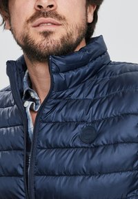 Chaleco acolchado azul marino con cuello alto, textura de acolchado y un parche de logo negro. Frontal con cremallera y superpuesto con una camisa a cuadros debajo.