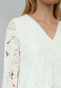 Blouse blanche en dentelle avec un décolleté en V, ornée de motifs floraux et de manches longues transparentes. Le tissu possède une texture délicate et des détails raffinés.