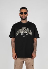 Čierne bavlnené tričko s voľným strihom a krátkymi rukávmi. Na prednej strane sa nachádza odvážna, štýlizovaná grafika "MJGONZALES" v smotanovej a zlatej farbe.