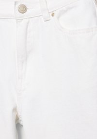 Gros plan sur un jean en denim blanc montrant la ceinture, le bouton en métal, la boucle de ceinture, la poche avant et les détails des coutures.