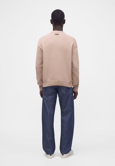 Homme debout, dos tourné, portant un pull beige, un jean bleu ample et des baskets blanches sur un fond blanc uni.