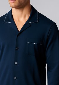Camicia da pigiama blu navy con colletto a fattura, tasca frontale e dettagli in piping bianco. Realizzata in un tessuto liscio e leggero con chiusura a bottoni.