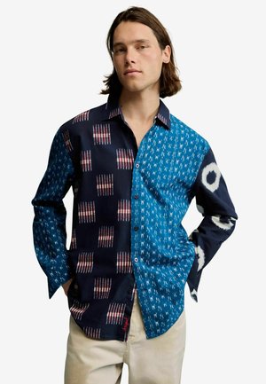 Mężczyzna ubrany w koszulę na guziki z długim rękawem w wzory patchworkowe w kolorach niebieskim, czarnym i białym, zestawioną z jasnobeżowymi spodniami.
