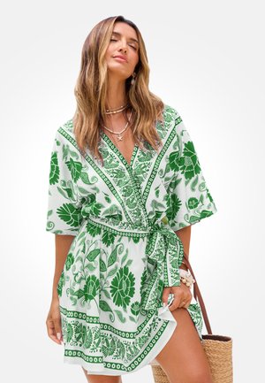 Kobieta w zielono-białej sukience kopertowej w kwiaty z wiązanym talią, trzymająca plecioną torbę, nosząca warstwowe naszyjniki i bransoletki.