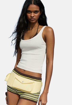 Fiatal nő hosszú sötét hajjal, fehér ujjatlan felsőt, csíkos kötött rövidnadrágot és sárga övtáskát visel egyszínű háttér előtt.