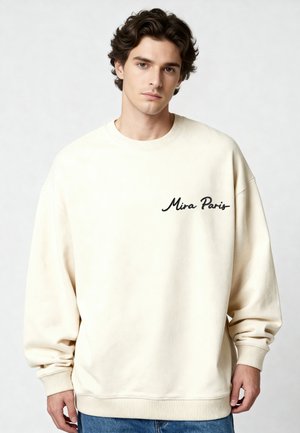 Jeune homme aux cheveux ondulés portant un sweat-shirt beige ample avec l'inscription « Mira Paris » et un jean bleu, debout devant un fond uni.