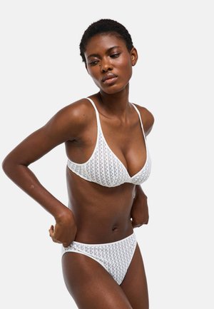 Femme portant un soutien-gorge blanc à motifs et une culotte assortie ajustant le vêtement du bas sur un fond uni.
