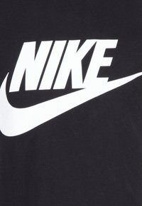 Juodas medvilnės marškinėlis su dideliu baltu Nike logotipu su švetimo dizainu, ryškiai centrine pozicija. Lygus paviršius, standartinė apvali apykaklė.