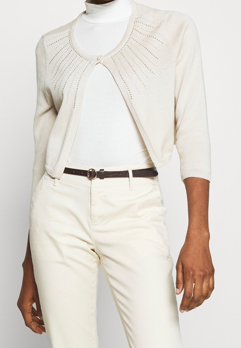 Femme portant un cardigan court crème avec des rayons cloutés sur un col roulé blanc, un pantalon taille haute crème, et une ceinture noire avec une boucle dorée.