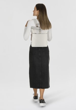 Zaino in beige chiaro, forma rettangolare con apertura superiore con zip, spallacci regolabili e superficie liscia. Indossato da una persona in una gonna nera.