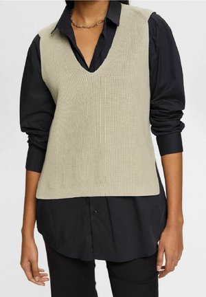Femme portant un gilet beige en maille sans manches par-dessus une chemise noire à manches longues aux manches retroussées, debout devant un fond uni.