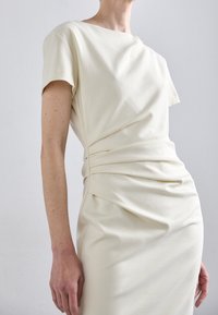 Krämfärgad klänning med korta ärmar, med rynkdetaljer i midjan och slät, texturerad tyg. Enkelt, elegant design.