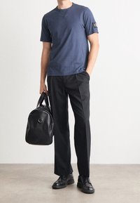 Mies, który ma na sobie granatową koszulę z krótkim rękawem, czarne spodnie, czarne skórzane buty i trzyma czarną skórzaną torbę w jednej ręce.