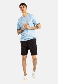 Camiseta de algodón azul claro, pantalones cortos cargo negros y zapatillas blancas. El modelo está de pie, con las manos juntas, frente a un fondo neutro.