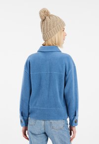 Veste en polaire bleue avec une coupe légèrement surdimensionnée, dotée d'un col et de poignets à boutons. Portée avec un bonnet en tricot beige avec un pompon.