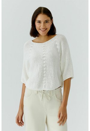 Lächelnde Frau mit schulterlangem braunem Haar, die ein locker sitzendes weißes Strickoberteil und hellbeige Hosen mit Kordelzug trägt.