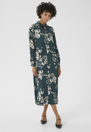 La modella indossa un vestito floreale color turchese con colletto, che presenta una vestibilità comoda. Le scarpe col tacco nere completano l'outfit.