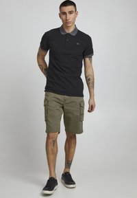 Zwart poloshirt met grijze kraag, kaki cargoshort en zwarte sneakers. Met zichtbare tatoeages op de armen. Neutrale achtergrond.