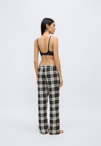 Kvinna med långt mörkt hår som är vänd åt sidan, bär en svart behå och svartvita rutiga vida byxor, står barfota mot en enkel bakgrund.