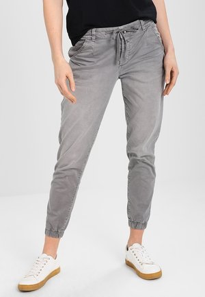 Kobieta w jasnoszarych spodniach dresowych z sznurkiem i białych trampkach stojąca na tle jednolitego jasnoszarego tła.