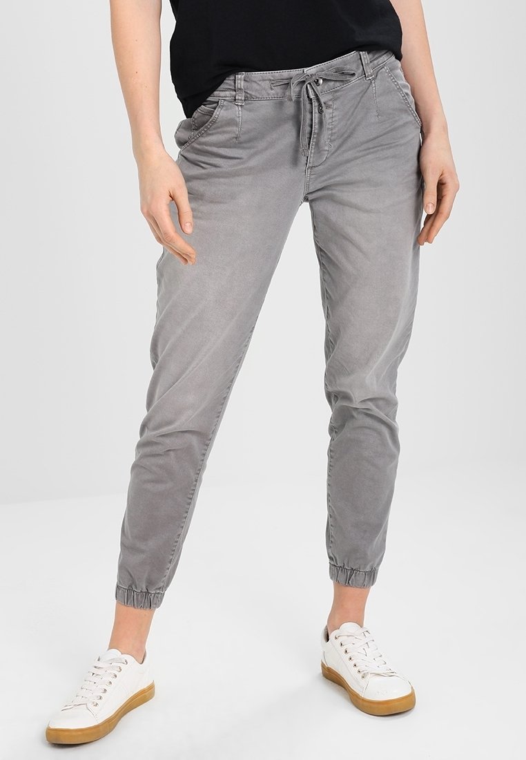 Femme portant un pantalon de jogging gris clair à cordon et des baskets blanches, debout devant un fond gris clair uni.