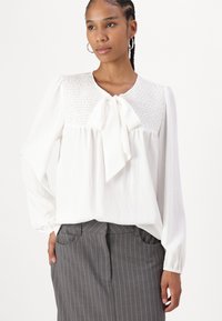 Blusa bianca con una parte superiore arricciata e testurizzata, caratterizzata da un fiocco al collo e maniche lunghe e ampie. Abbinata a pantaloni grigi a righe.