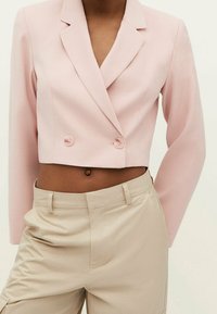 Jasnoróżowy krótki żakiet o kroju dwurzędowym, z wyciętym kołnierzem i ozdobnymi guzikami. Zestawiony z beżowymi spodniami o gładkiej fakturze.