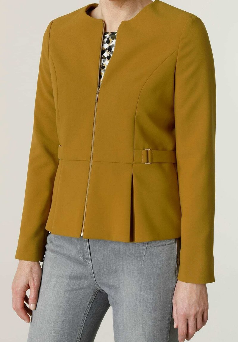 Femme portant une veste zippée jaune moutarde avec un détail de boucle sur le côté et un jean gris clair, debout devant un fond uni.