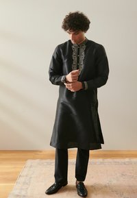 Czarny kurta z misternym srebrnym haftem wzdłuż kołnierza i rękawów, w połączeniu z czarnymi spodniami i błyszczącymi butami na beżowym dywaniku.