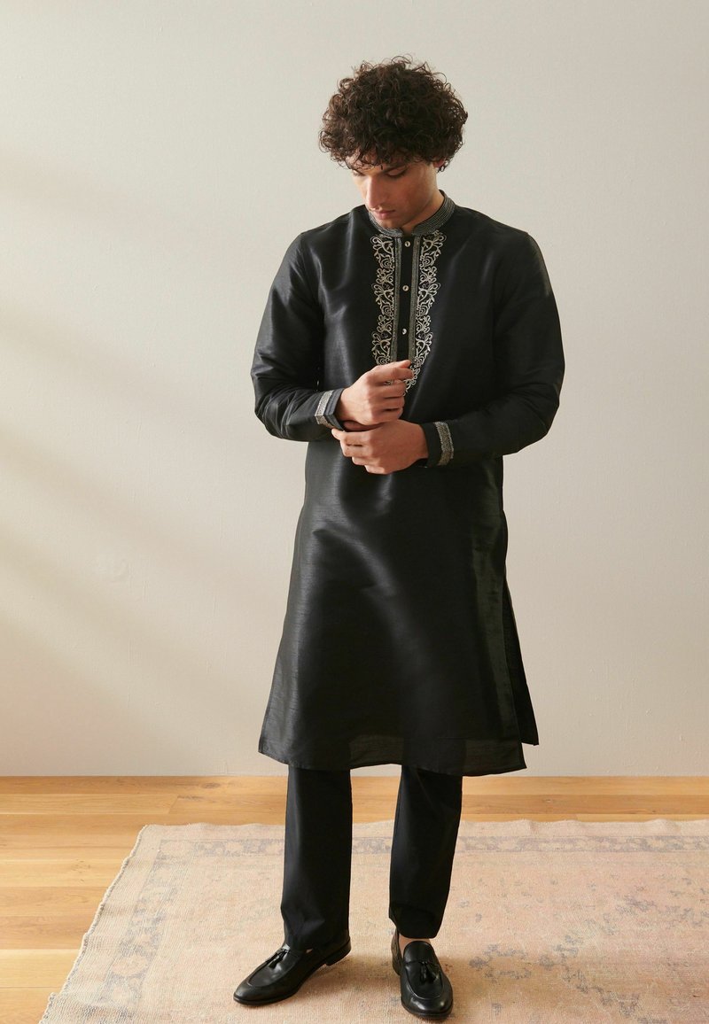 Musta kurta dengan bordir perak yang rumit di sepanjang kerah dan lengan, dipadukan dengan celana hitam dan sepatu mengkilap di atas karpet beige.