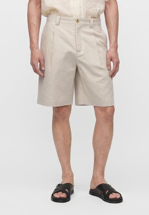 Shorts plissés beige portés avec une chemise crème et des sandales noires à brides croisées sur une personne debout devant un fond uni.