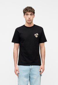 Νεαρός άνδρας στέκεται φορώντας μαύρο μπλουζάκι με μικρό μπεζ σχέδιο φοίνικα και ανοιχτό μπλε τζιν, μπροστά σε απλό λευκό φόντο.