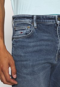 Närbild av en person som bär blå denimjeans med en liten logotyp på myntfickan och en ljusblå skjorta instoppad.