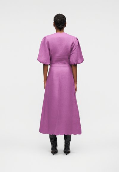 Femme aux cheveux tressés portant une robe midi violette à manches bouffantes et ourlet festonné, associée à des bottes à talons noires, vue de dos.