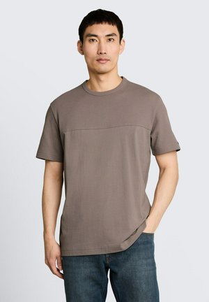 Mann trägt ein kurzärmeliges taupefarbenes Hemd mit strukturiertem oberen Bereich und blaue Jeans, steht mit einer Hand in der Tasche vor einem schlichten Hintergrund.