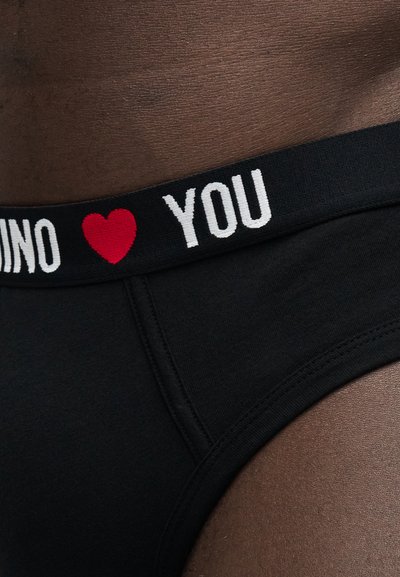 Κοντινή λήψη μαύρης ζώνης εσώρουχου σε σκούρο δέρμα με άσπρο κείμενο "MOSCHINO" και "YOU", χωρισμένα με ένα κόκκινο σύμβολο καρδιάς.