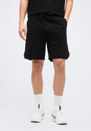 Männlicher trägt schwarze Shorts mit Kordelzug, weiße Wadenhochsocken und weiße Sportschuhe und steht vor einem einfarbigen Hintergrund.