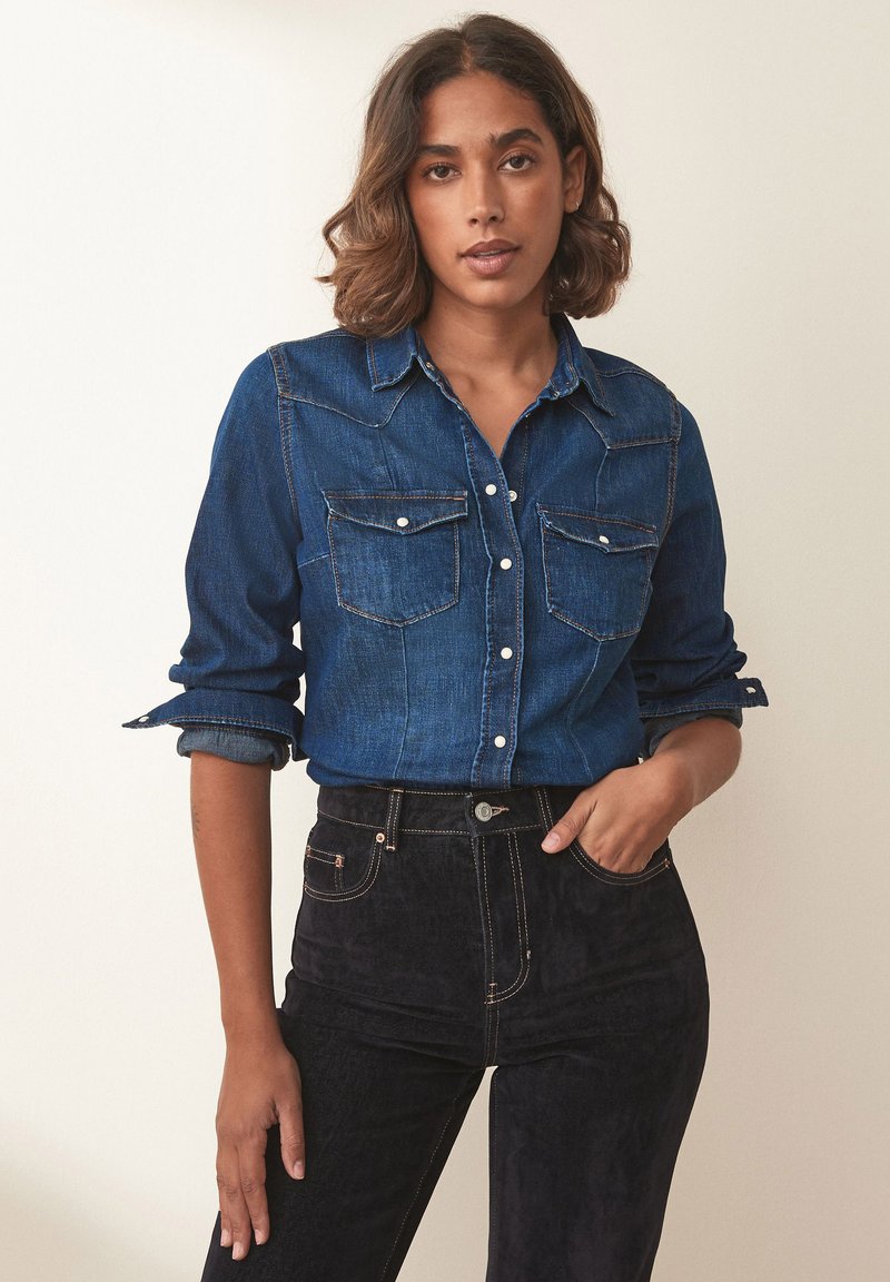 Cămașă button-up din denim închis, cu două buzunare frontale, mâneci suflecate și detalii subtile de cusătură, asortată cu pantaloni negri cu talie înaltă.