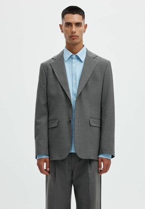 Jeune homme aux cheveux courts portant un costume gris à fines rayures et une chemise bleu clair à boutons, debout devant un fond clair uni.