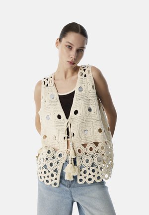 Crocheted vest in cream with circular cutouts and metallic accents. Features a tie closure and tassel detail at the bottom.