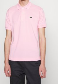 Camisa polo de color rosa claro, hecha de algodón, con cuello extendido, mangas cortas y logo bordado de Lacoste en el lado izquierdo del pecho.