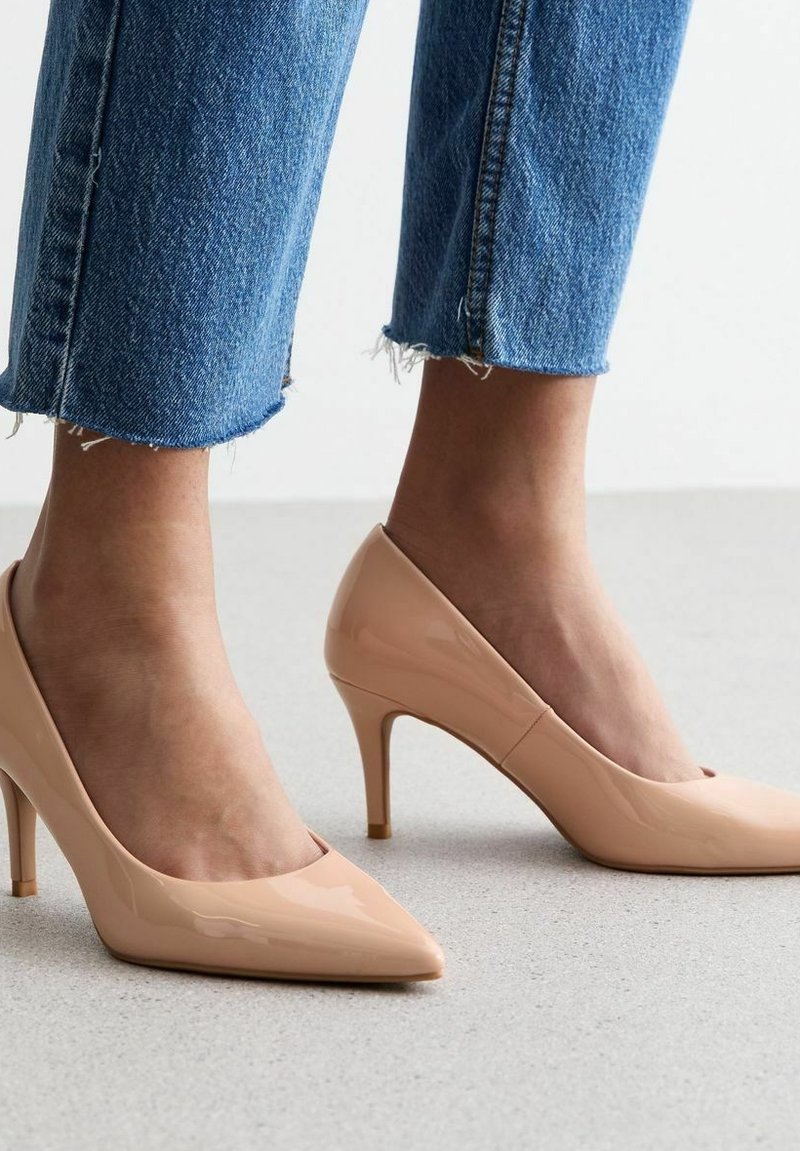 Zapatos de tacón alto de charol beige con punta afilada y tacón delgado. Combinados con jeans de mezclilla azules rasgados, mostrando un diseño elegante.